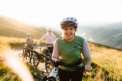 3 Frauen mit ihren Mountainbikes auf einem Trail umgeben von Almwiesen und Bergen 