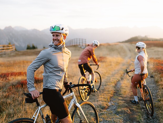 3 Personen auf Gravel Bikes auf einer Schotterstraße, Ringsum umgeben von Almwiesen und Berge 