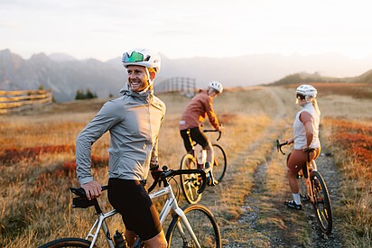 3 Personen auf Gravel Bikes auf einer Schotterstraße, Ringsum umgeben von Almwiesen und Berge 