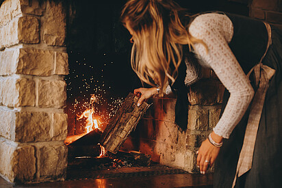 A woman by the fireplace is adding wood to the burning stove.
